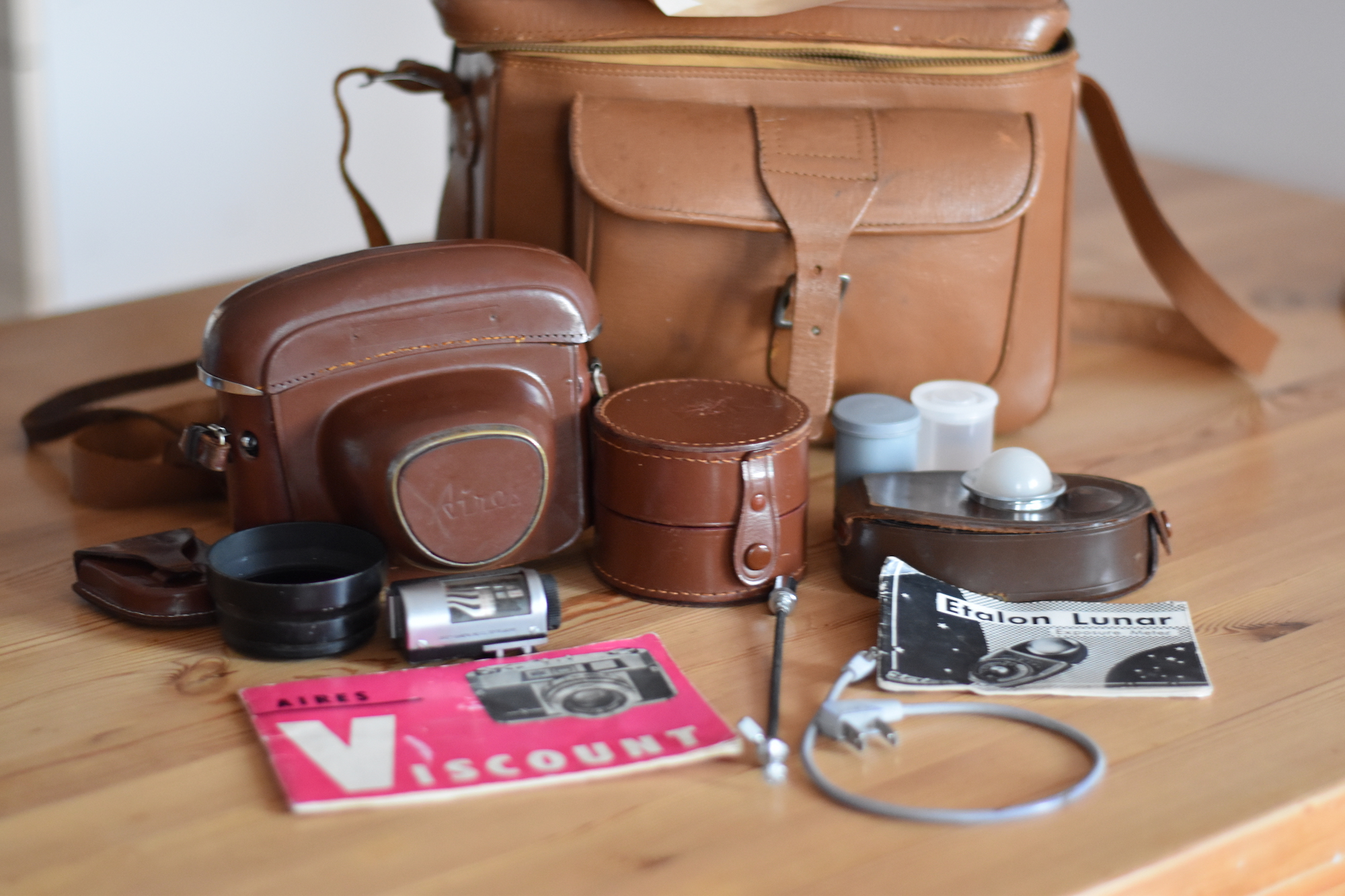 A group of camera equipment in front of a leather camera bag. Including a camera in a full leather case, a leather lens box, a light meter, some film canisters, and a manual