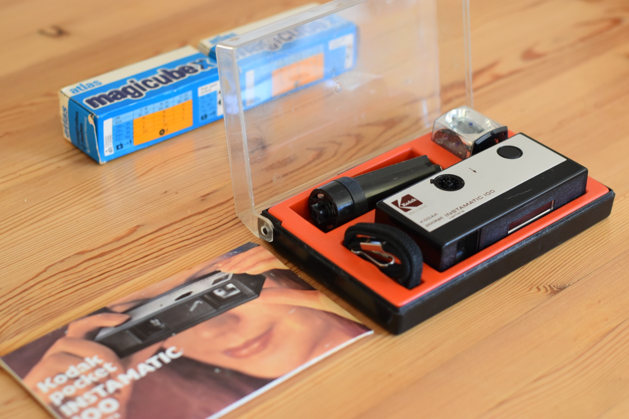 Kodak Instamatic Pocket 100 in original plastic box. Box top is clear, box base is black with a red plastic insert that holds the camera components. Manual is on table in front of box, has a picture of a young woman holding the camera up to her eye