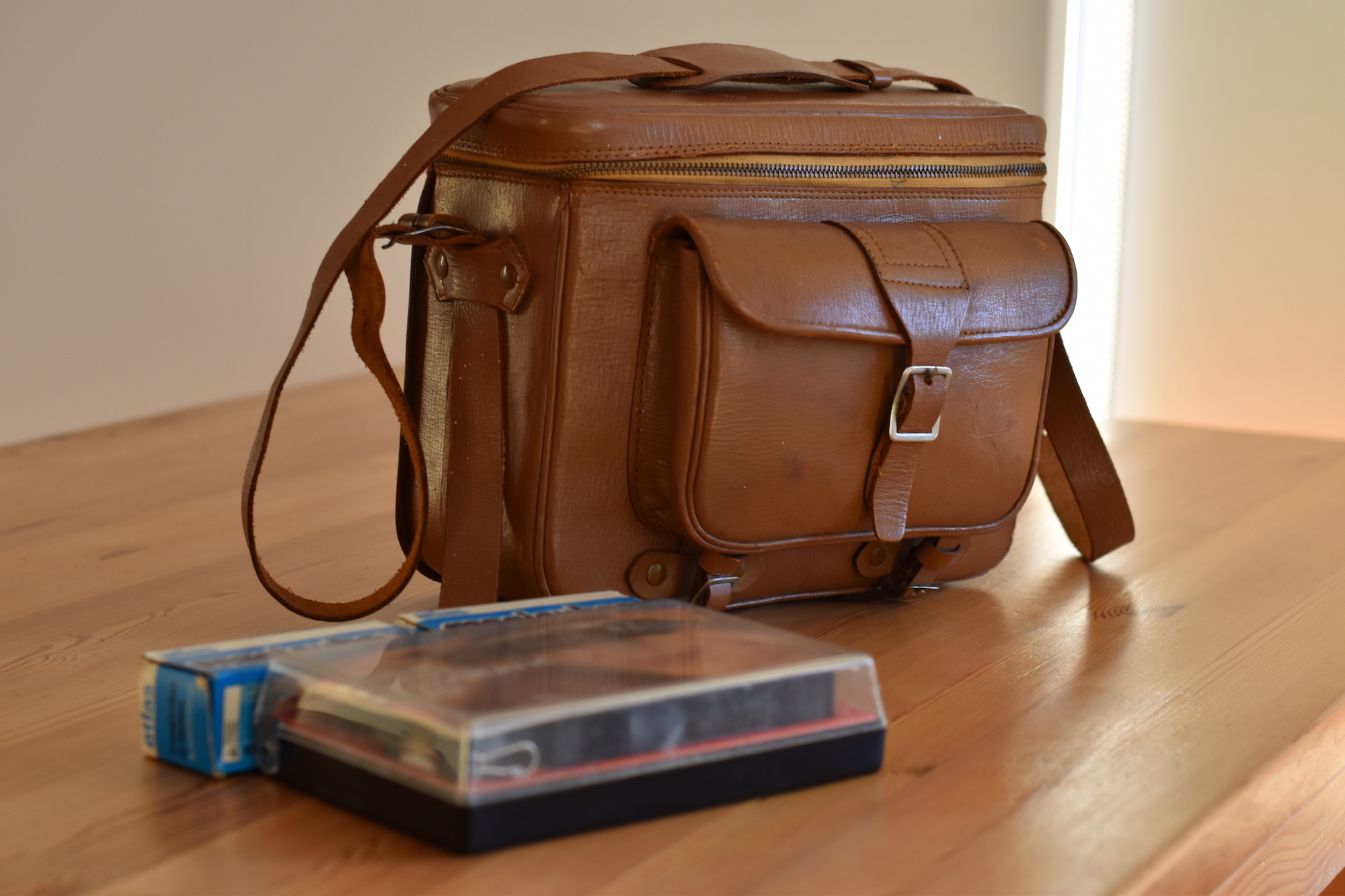 Kodak Instamatic Pocket 100 in original plastic box in front of a light brown leather camera bag with zip-around top and front buckled pocket